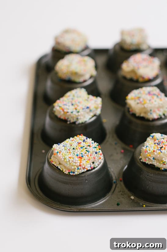 Ingredients laid out for making Ice Cream Cookie Cups, including pre-made cookie dough, colorful sprinkles, a tub of ice cream, and a muffin tin.