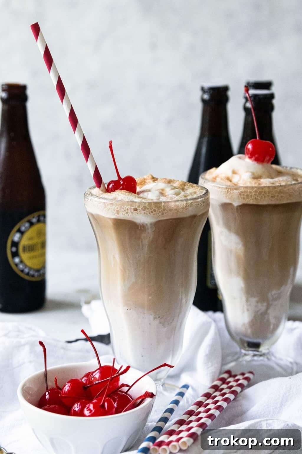 Two tall glasses of vanilla root beer floats, topped with whipped cream and cherries, photographed from an appealing angle.