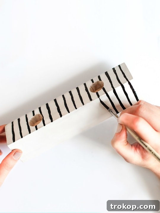 Handmade Patterned Candle Decor 7 A hand meticulously painting black grid lines onto a white-sprayed wooden candle holder with a small brush, showcasing the pattern application step in a DIY candle holder project.