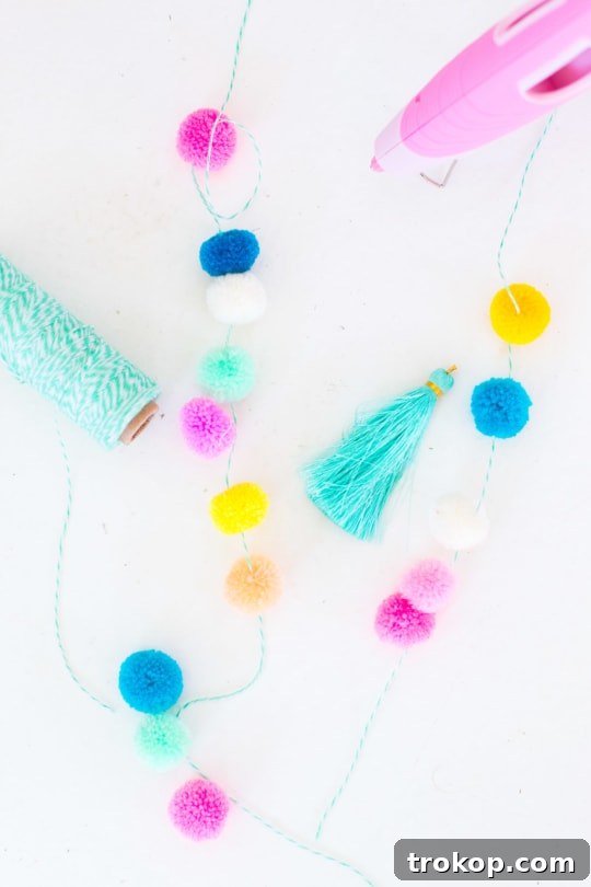 Detail of threading colorful pom poms onto twine to create a garland, showing the secure knotting process.