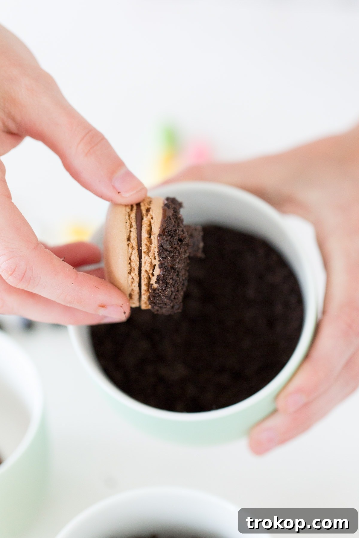 A hand dipping a chocolate macaron into melted chocolate.