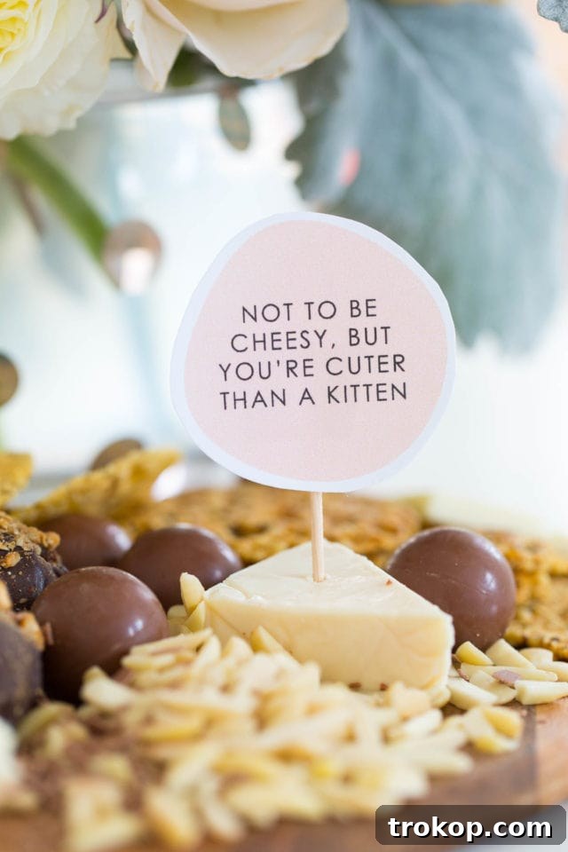 Close-up of a pink cheese board marker with a witty message, 'You're Grate!', nestled among cheese, grapes, and crackers on a rustic wooden board.