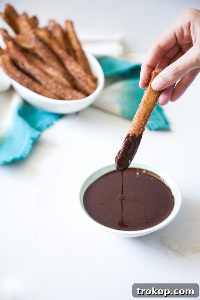 A close-up of freshly fried cinnamon churros stacked next to a small bowl of dark, glistening chili chocolate tequila dipping sauce.