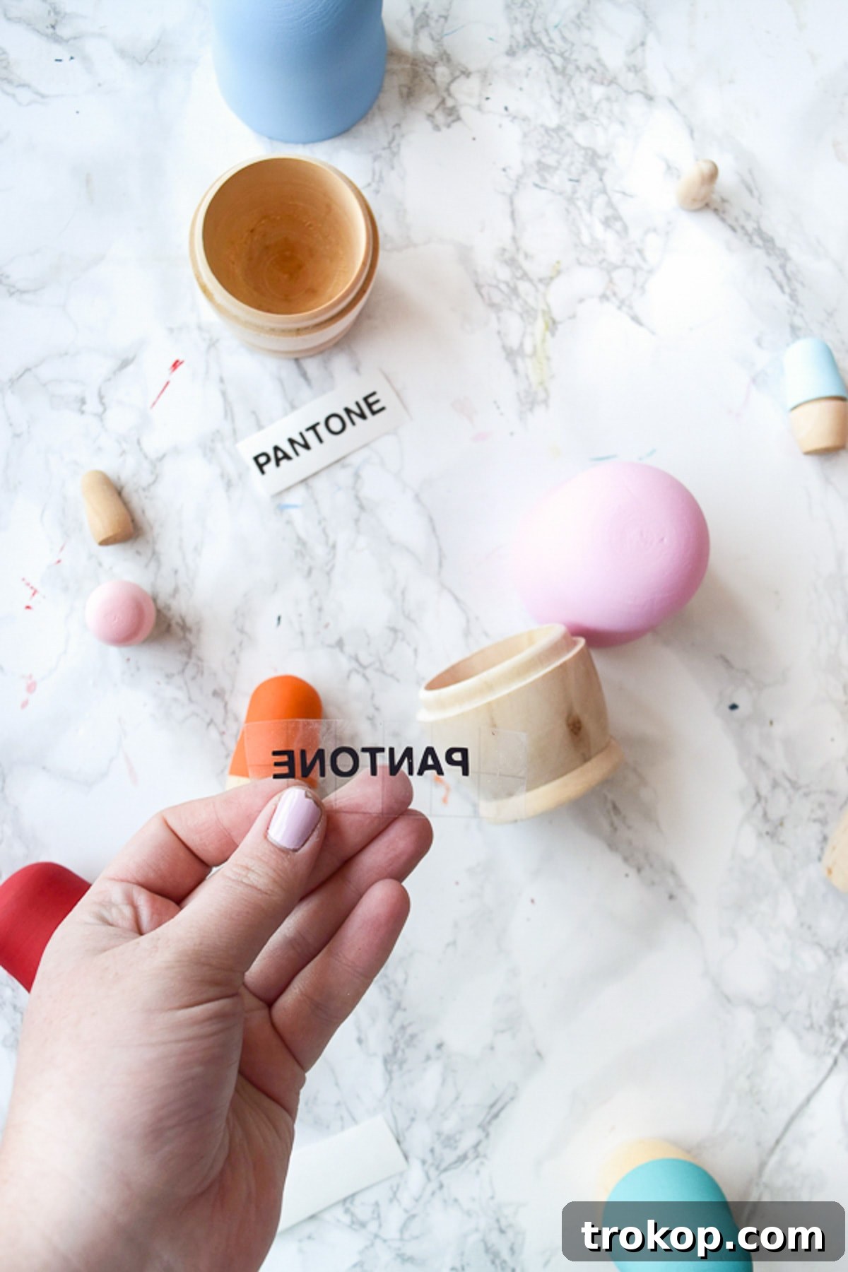Applying custom-cut 'PANTONE' vinyl lettering using transfer tape to a freshly painted wooden nesting doll lid, ensuring precise and clean typography.