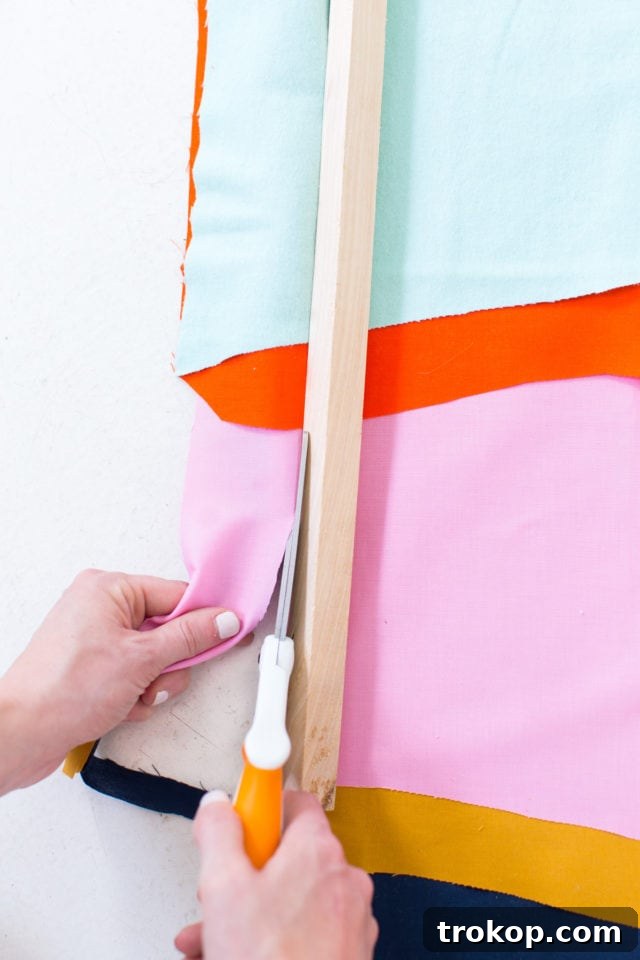 Close-up of excess fabric being trimmed from the top of a wooden dowel rod, finalizing the edge of the wall art