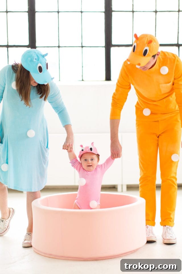 Our Handmade Hungry Hippo Crew 2 A family dressed in vibrant DIY Hungry Hungry Hippos costumes, featuring a baby in a pink hippo costume within a custom-designed baby walker that resembles the board game, surrounded by white ball pit balls. The scene captures the joy and creativity of a unique family Halloween costume idea.