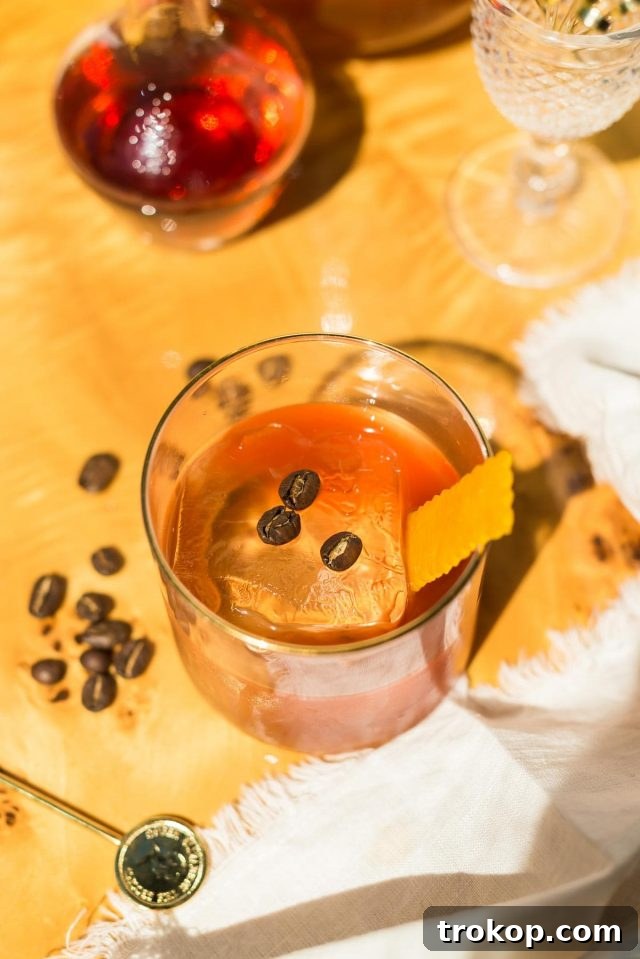 A hand holding a Coffee Old Fashioned cocktail garnished with orange peel and coffee beans.