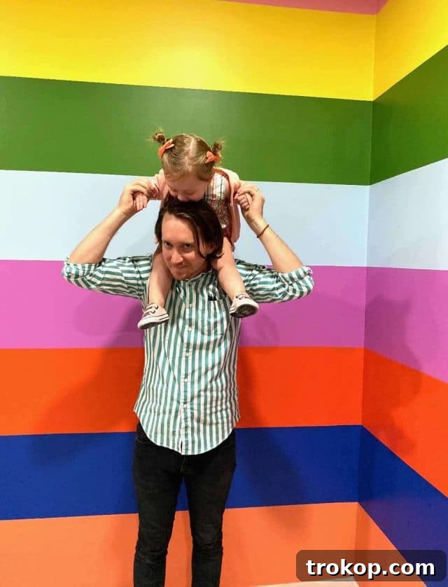 A woman riding on a man's back, both smiling, surrounded by colorful art installations at the Color Factory, symbolizing joy and lightheartedness amidst the world's complexities.