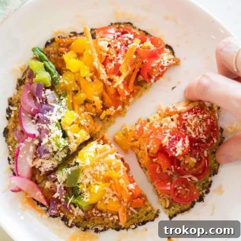 Overhead view of a vibrant rainbow veggie-topped pizza with a cauliflower crust and a hand reaching for a slice