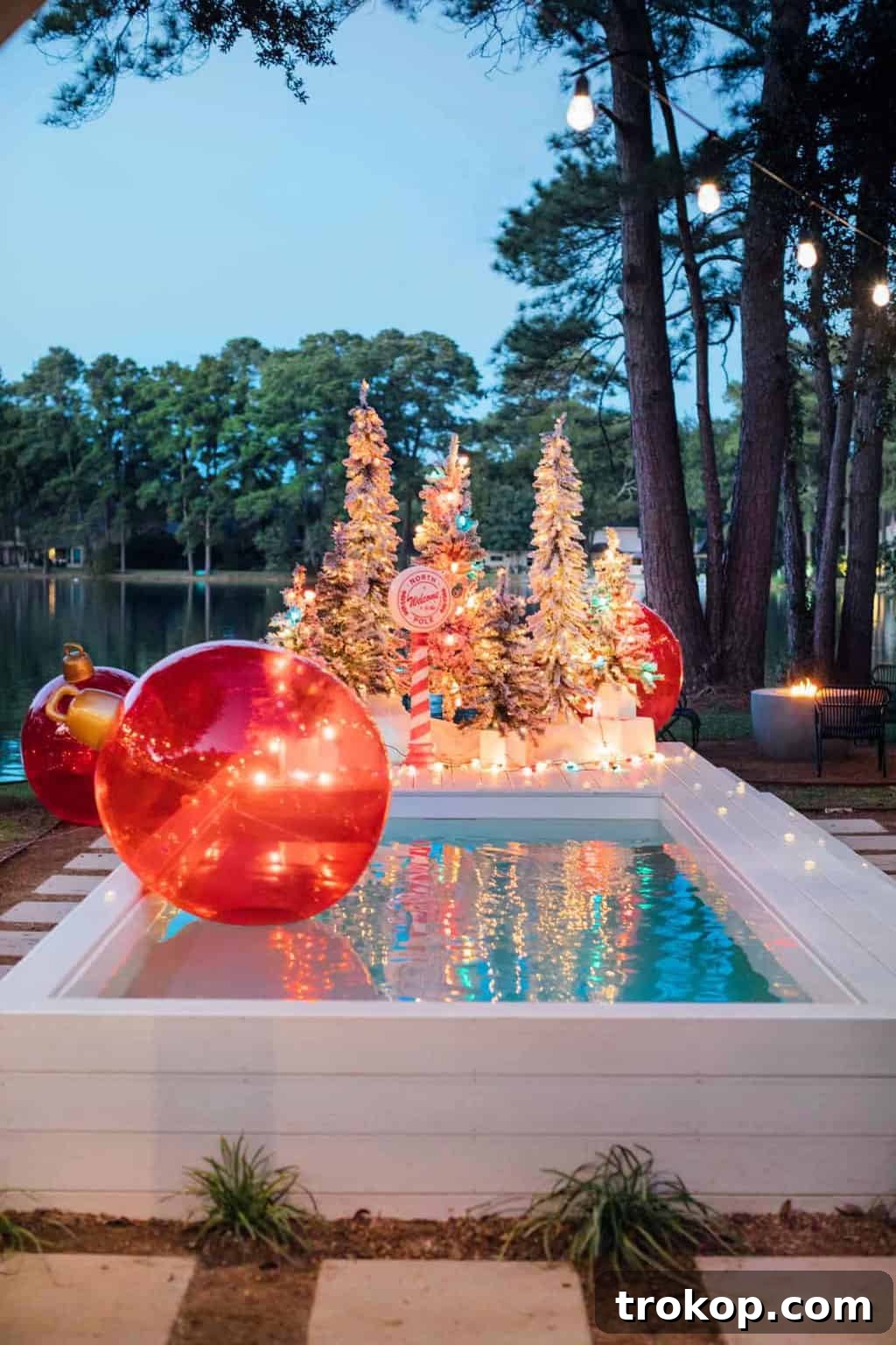 Santa's Poolside Paradise Christmas 12 A whimsical outdoor Christmas decoration featuring a jumbo red ornament nestled among flocked Christmas trees and ceramic lights.