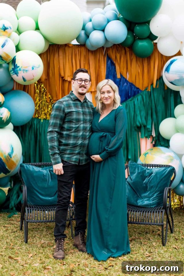 A husband and wife posing affectionately in front of their festive drive-thru shower backdrop, surrounded by balloons and giant 'OH BABY' marquee letters