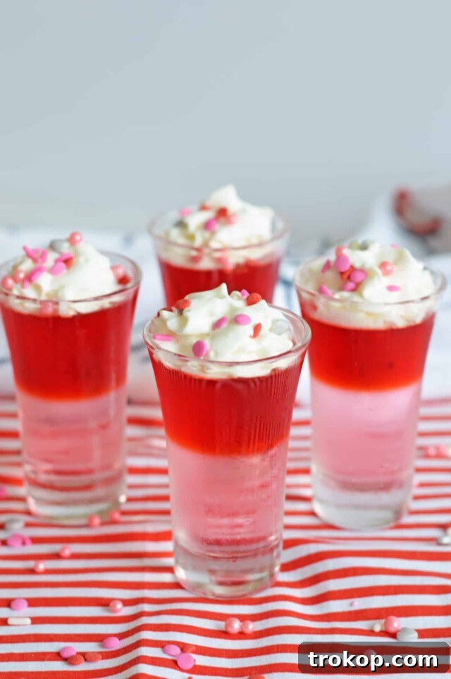 Valentine’s Day Jello Shots featuring layers of pink and red jello, garnished with whipped cream and sprinkles.