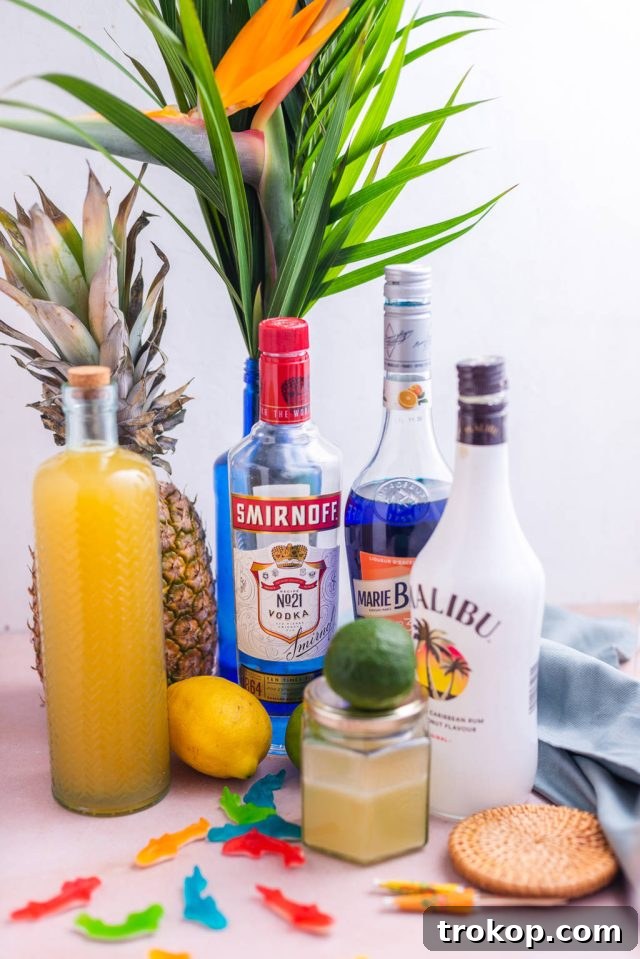 A flat lay image displaying all the vibrant ingredients laid out for making the Fish Bowl Drink, including bottles of liquor, pineapple juice, blue curaçao, and a bowl of Swedish Fish candies.