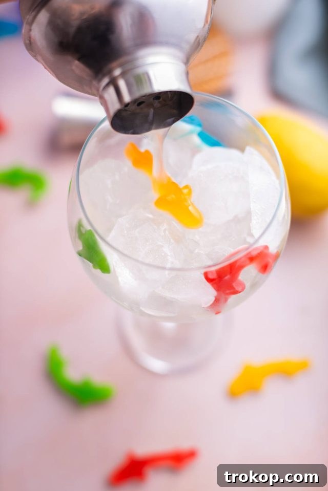 Pouring the vibrantly blue mixed cocktail from a shaker into a prepared balloon glass filled with ice and Swedish Fish, completing the Fish Bowl Drink.