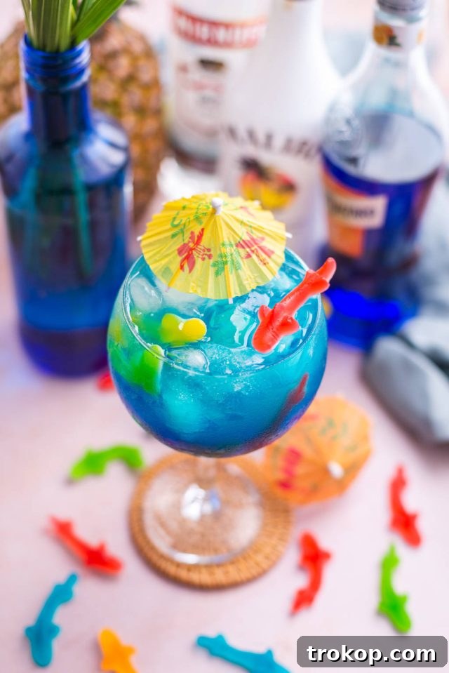 A single Fish Bowl Drink in a large glass, garnished with Swedish Fish and an umbrella, showcasing its vibrant blue color and inviting appeal.