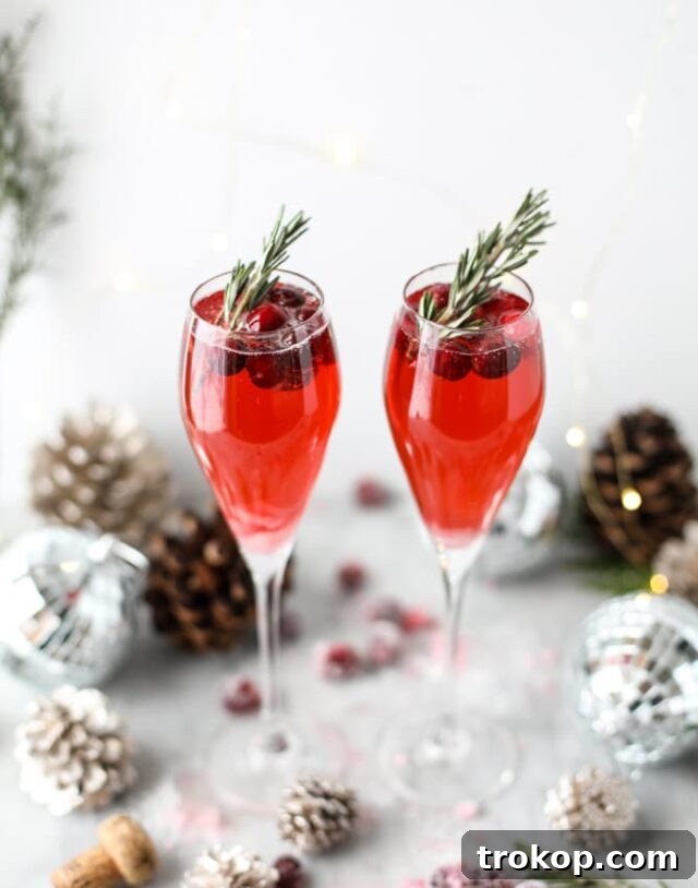 25 Easy & Festive Christmas Cocktails for Holiday Merriment 10 Christmas Morning Mimosa for Christmas cocktail recipes, a bubbly red drink in a champagne flute with cranberries and rosemary.