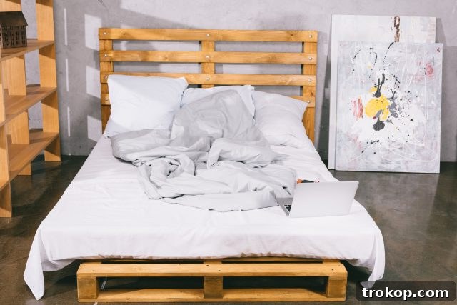 Laptop resting on a small, rustic wooden table made from pallets next to a bed in a cozy bedroom setting, highlighting versatile pallet furniture